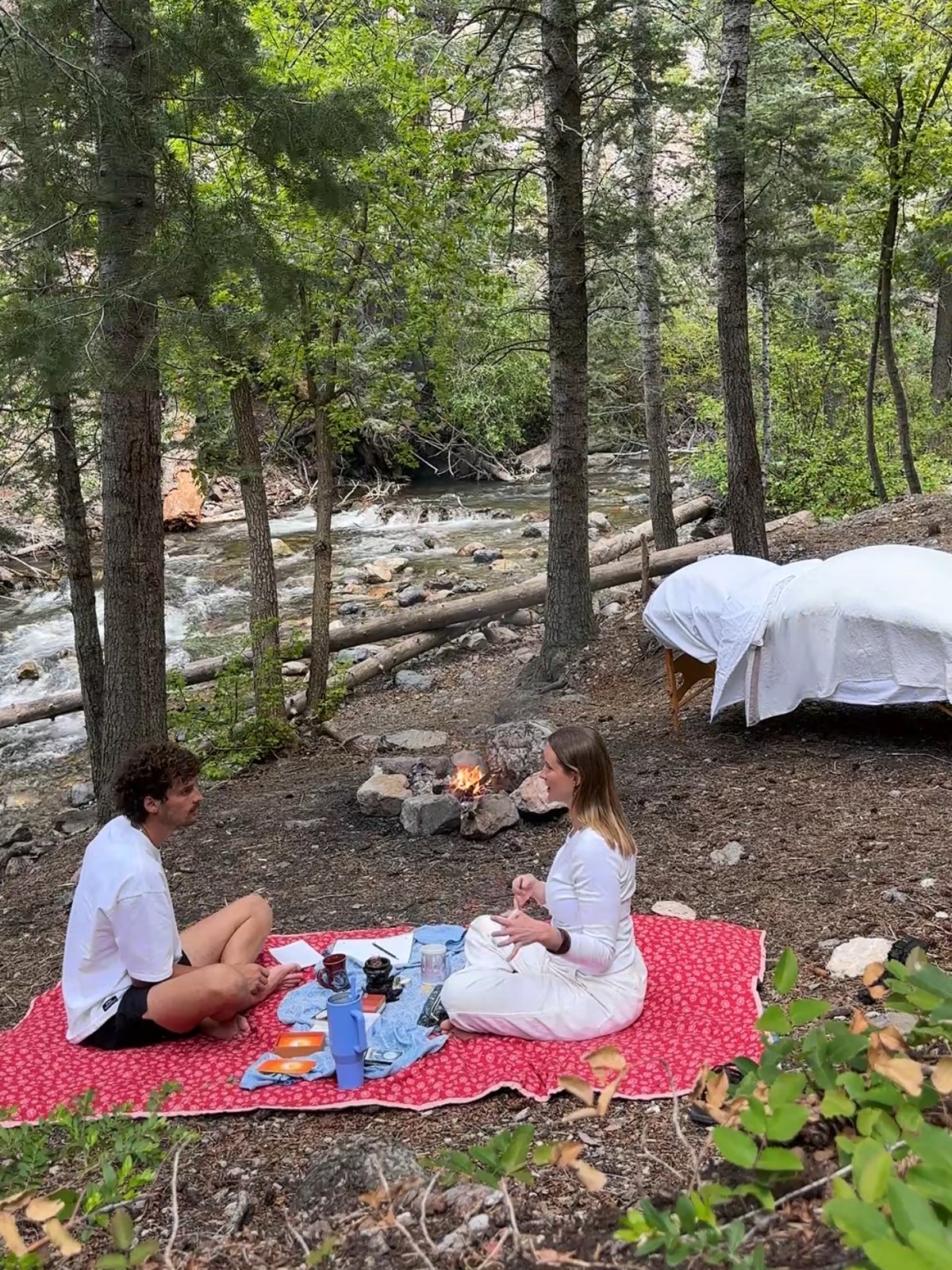 Two people sitting in conversation on a red blanket beside a creek and a small fire