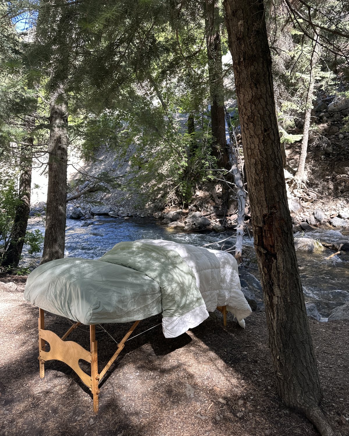 A treatment table dressed in a soft duvet beneath tall pines, beside a mountain creek